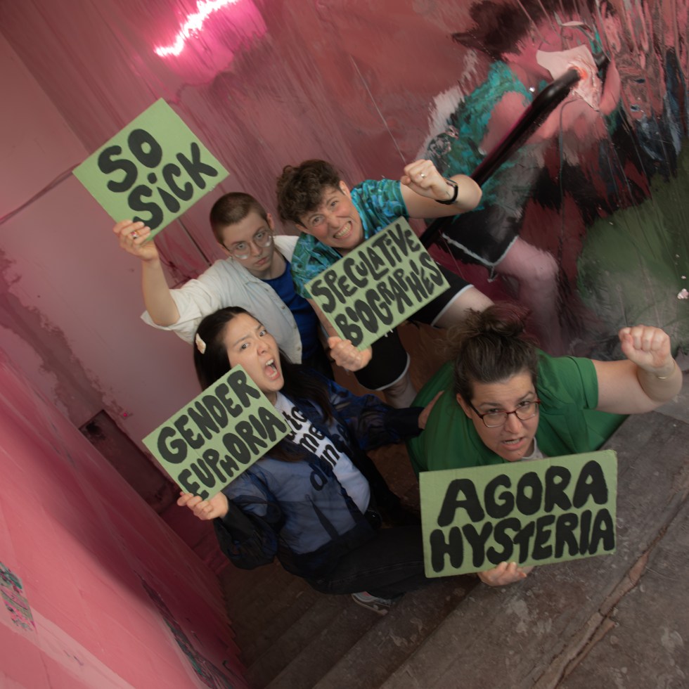 Hyunsin is holding the sign "Gender Euphoria", Luca is holding "So Sick", Jones is holding "speculative biographies" and Elena is holding the sign with "Agora Hysteria". Everyone is making a face as if they are shouting